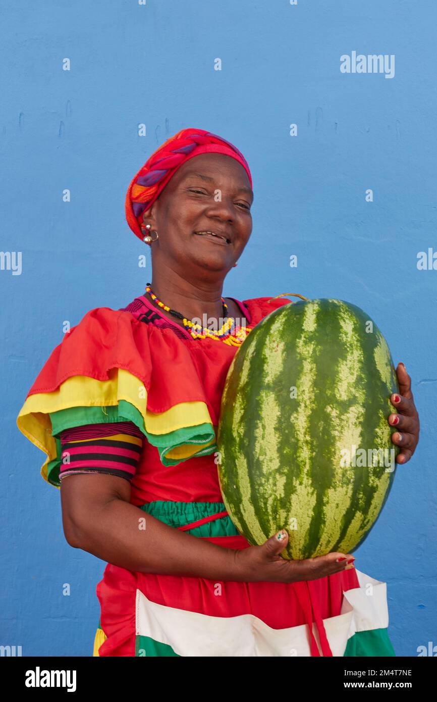 A vertical footage of a Palenquera holding a big heavy watermelon, dressed with colorful clothes on a blue background in Cartagena, Spain Stock Photo