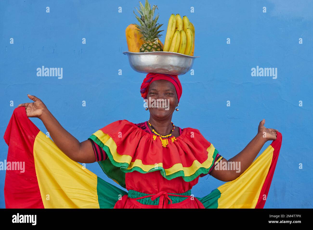 A regular footage of a Palenquera holding a big bowl of fruits above head, dressed with colorful clothes on a blue background in Cartagena, Spain Stock Photo