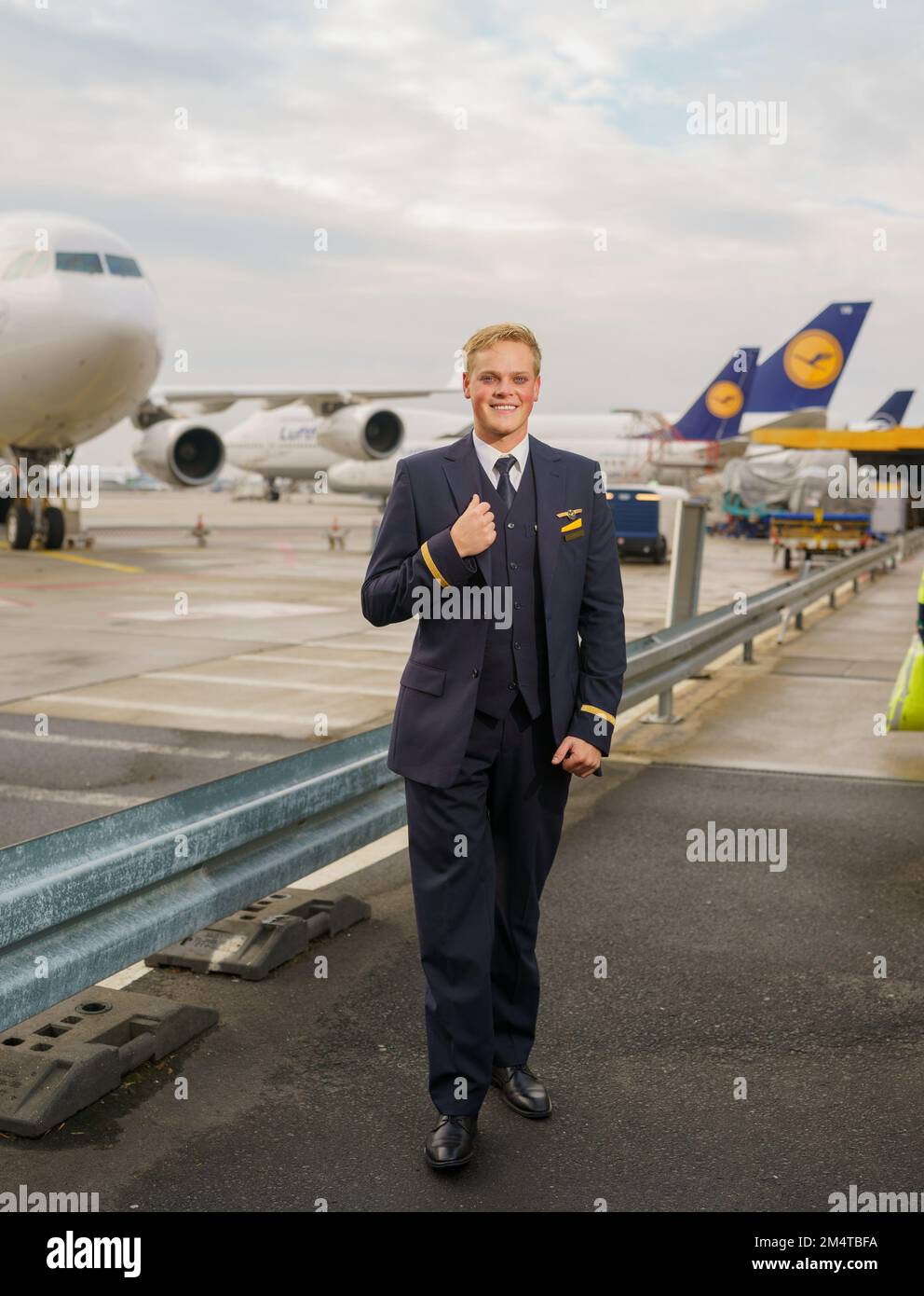 PRODUCTION - 08 December 2022, Hessen, Frankfurt/Main: Alexander Böhmer,  Lufthansa flight attendant, stands in front of several of his airline's  aircraft wearing his service uniform. The young flight attendant lost a leg