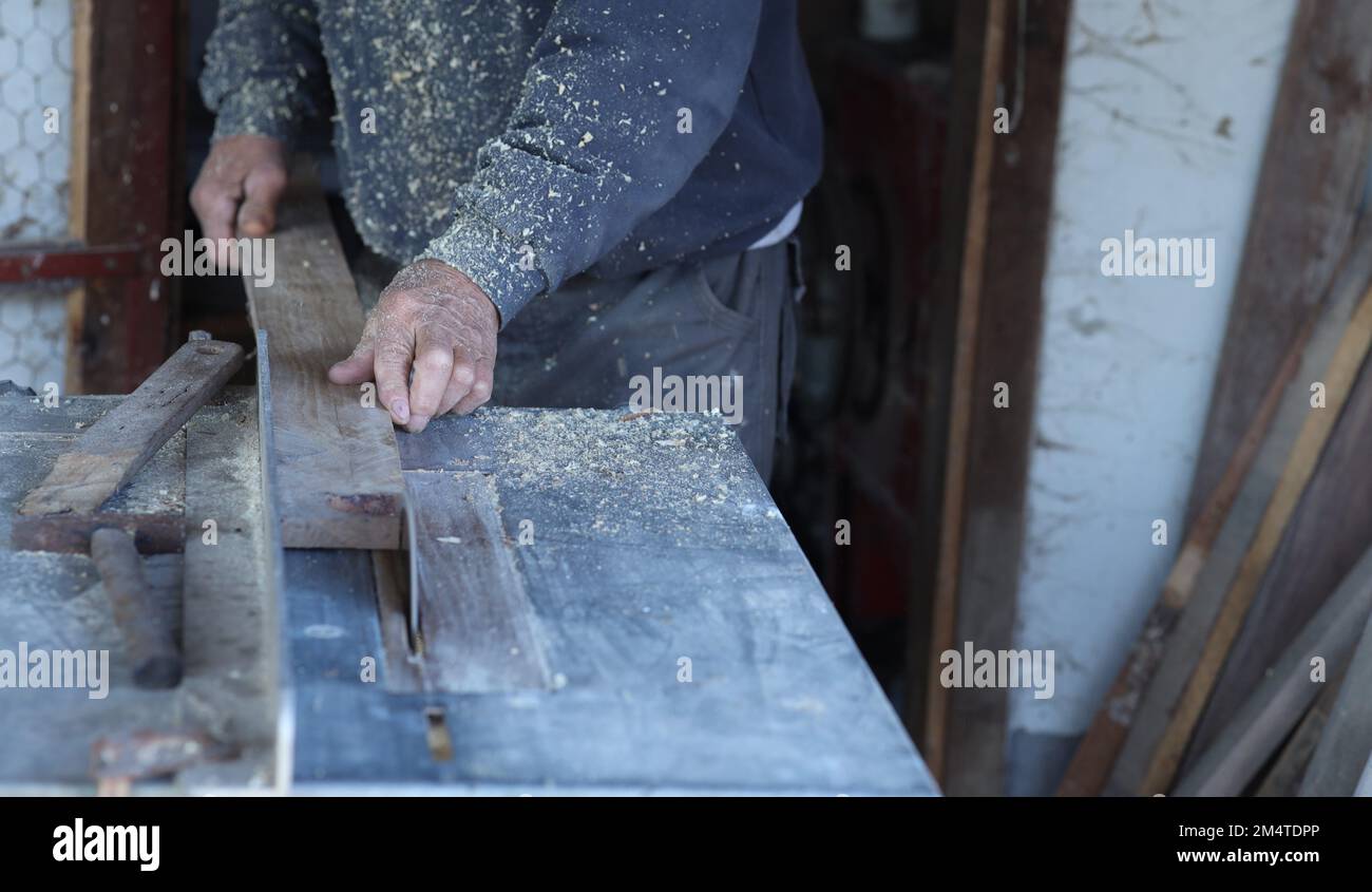 an elderly carpenter is cutting wood on a circular saw.close up photo of the hand holding and pushing the batten towards the saw. Stock Photo