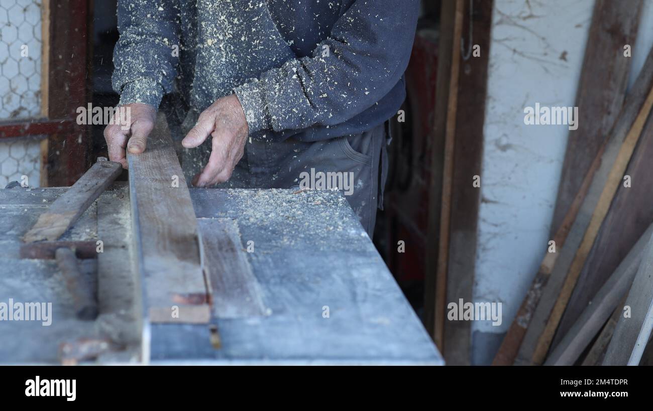 a carpenter processes wood on a circular saw.the focus is on the hands of the worker pushing the batten towards the circular saw. there is sawdust Stock Photo