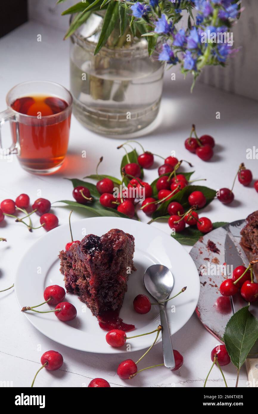 Composition on white background - delicious chocolate cake with sweet cherry, white plate with part of tart, cup of tea and glass jar with beautiful s Stock Photo