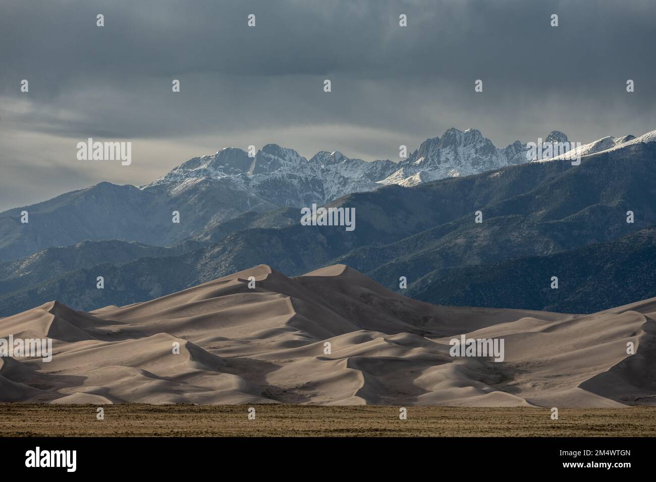 Drifts of Sand Dunes Form in the Wind Below Snow Capped Mountains in Great Sand Dunes National Park Stock Photo