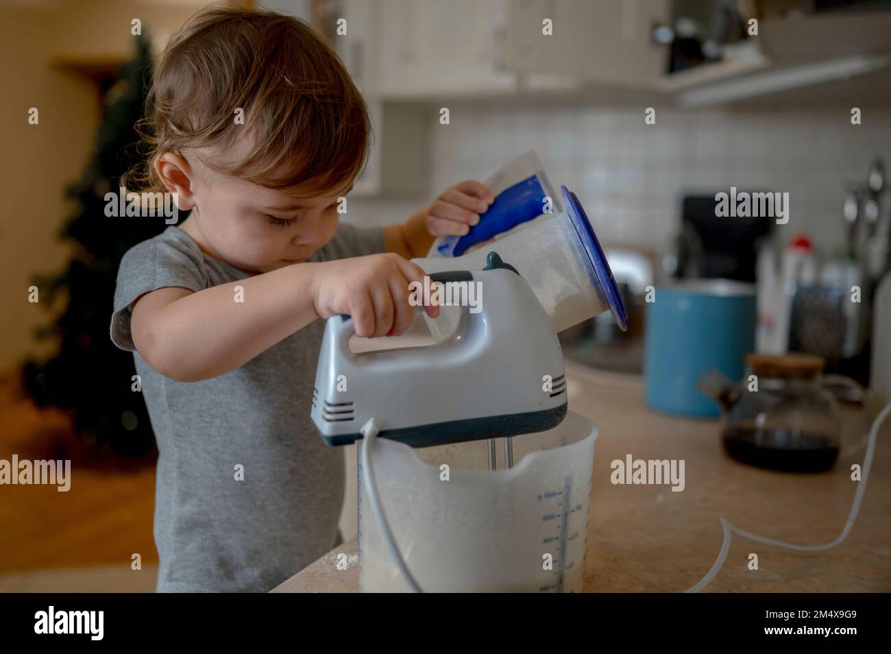 Baby boy pouring flour in container and mixing with electric mixer at