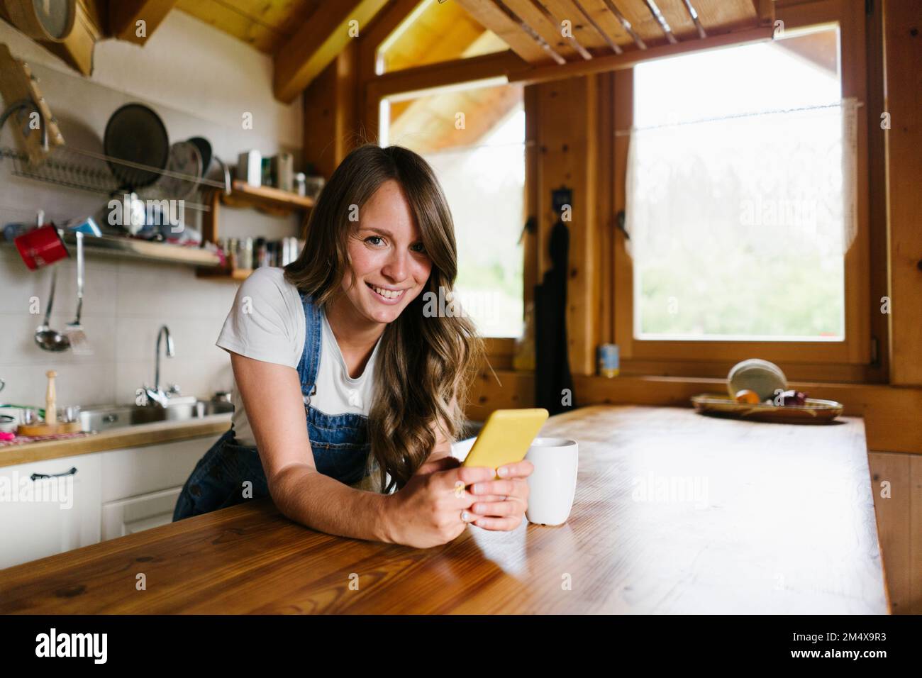 Smiling young woman leaning with smart phone on kitchen island Stock Photo
