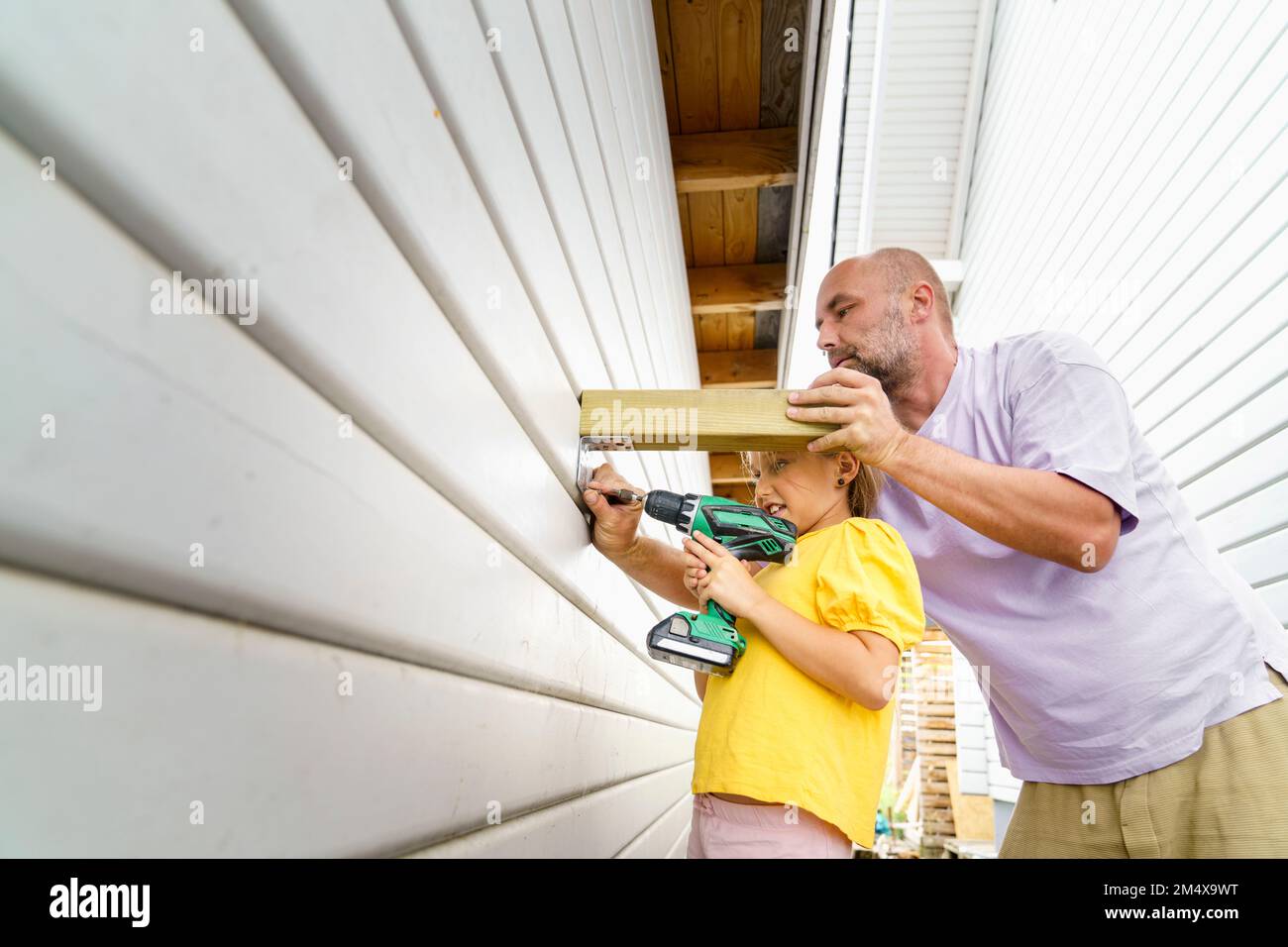 Father teaching daughter to build wooden wall using drill near house Stock Photo