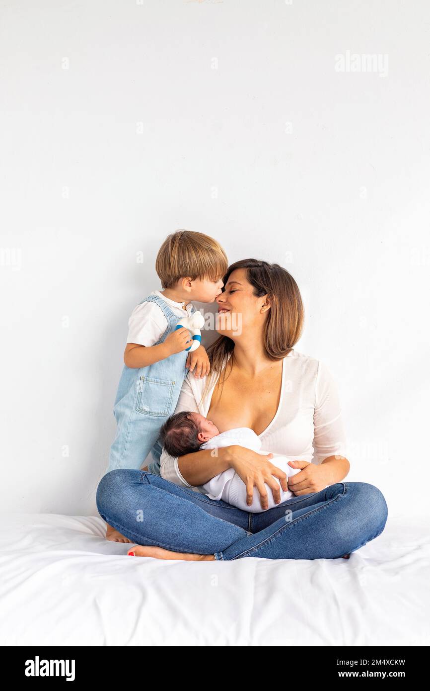 Son kissing mother breastfeeding daughter sitting on bed at home Stock Photo
