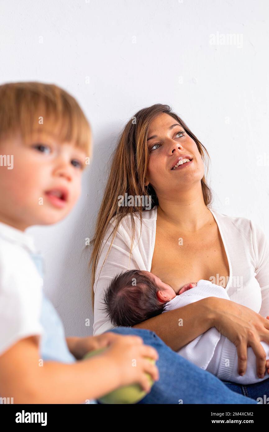 Contemplative mother breastfeeding newborn daughter by son at home