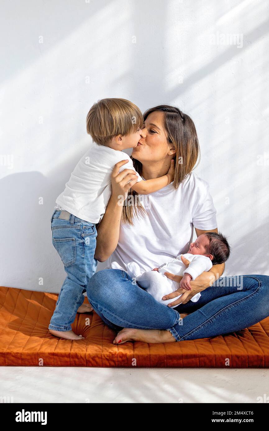 Son kissing mother sitting with newborn baby girl at home Stock Photo