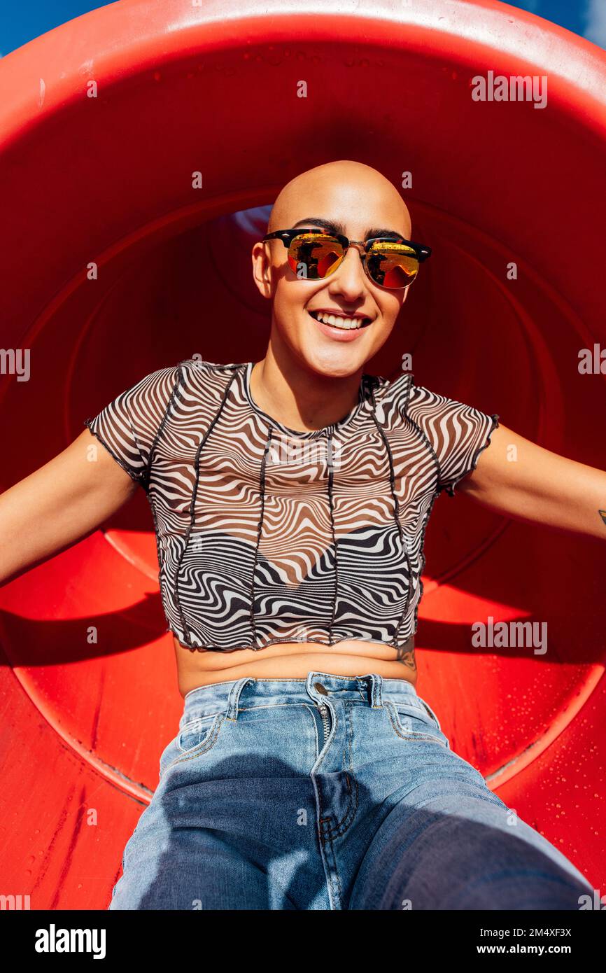 Happy woman with shaved head wearing sunglasses sitting in slide Stock Photo