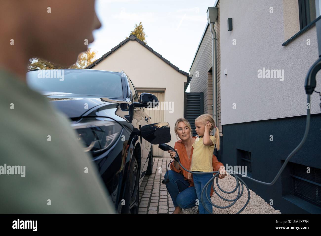 Woman holding electric car charger plug looking at daughter in front yard Stock Photo