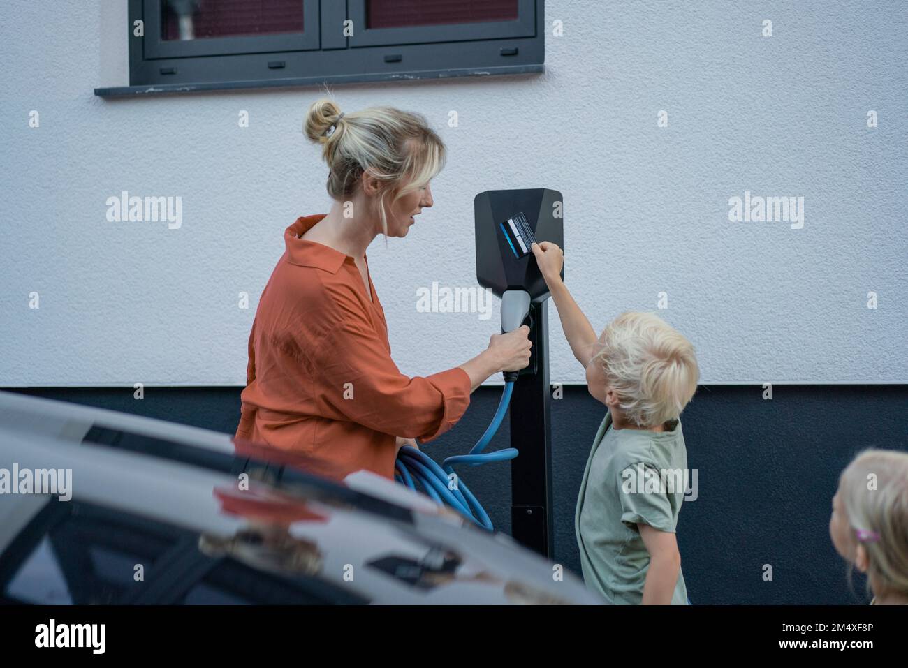 Boy paying through credit card by mother holding electric car charger plug in front yard Stock Photo