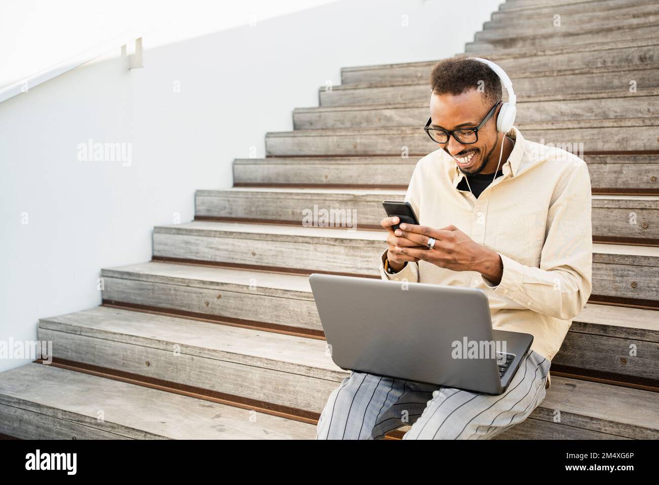 Happy freelancer text messaging through smart phone sitting with laptop on steps Stock Photo
