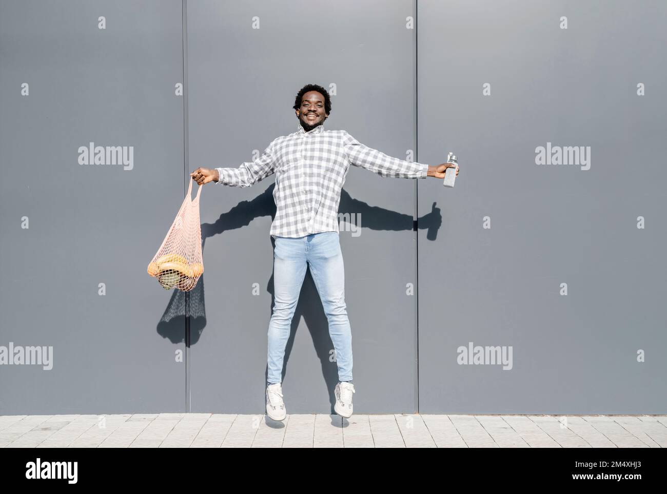 Cheerful jumping in front of gray wall holding mesh bag Stock Photo