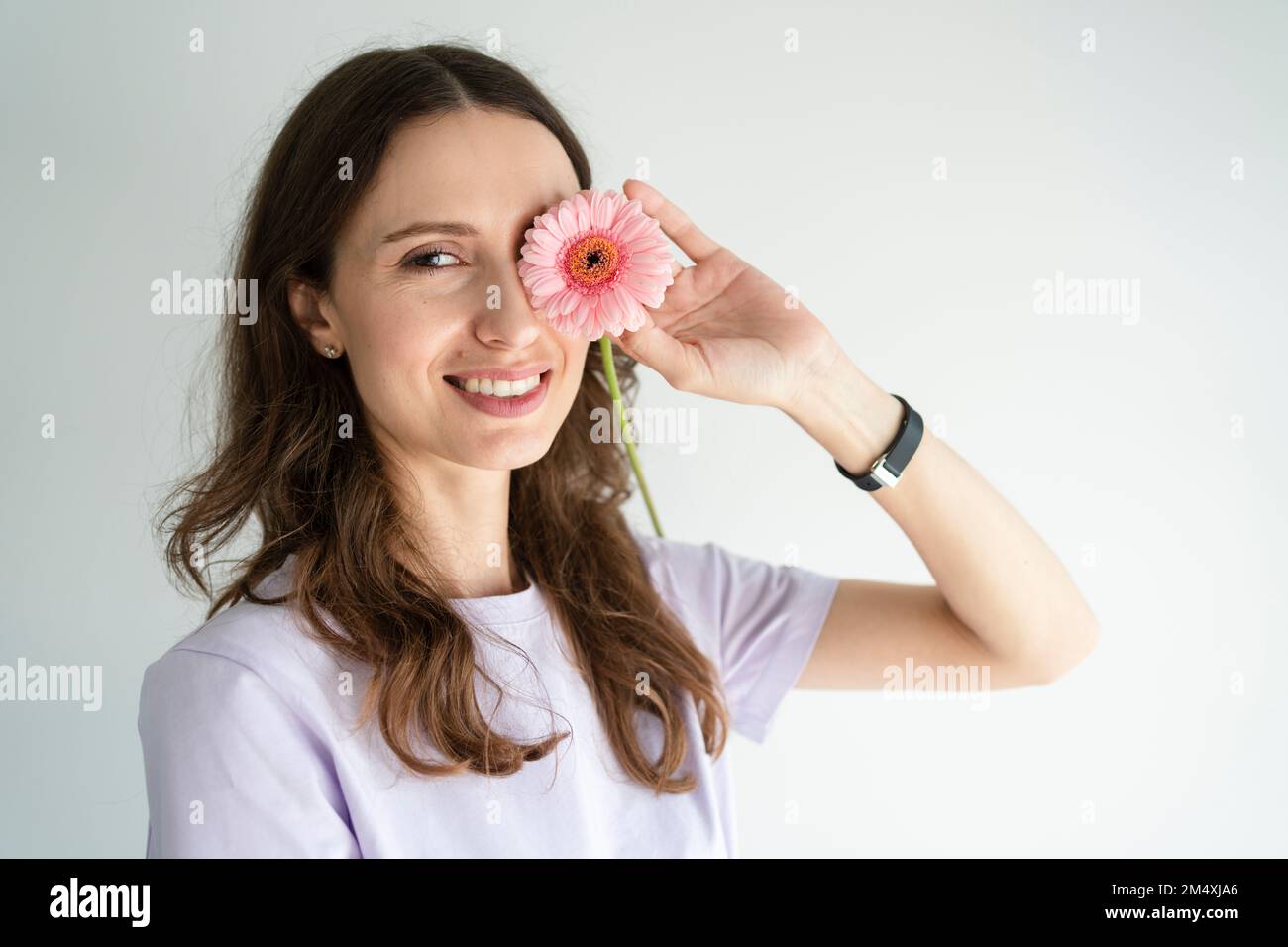 Happy woman covering eye with gerbera flower in front of white wall