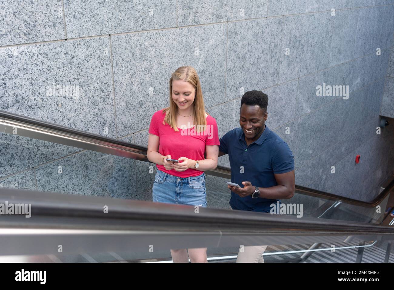 Smiling man and woman using smart phones moving up on escalator by wall Stock Photo