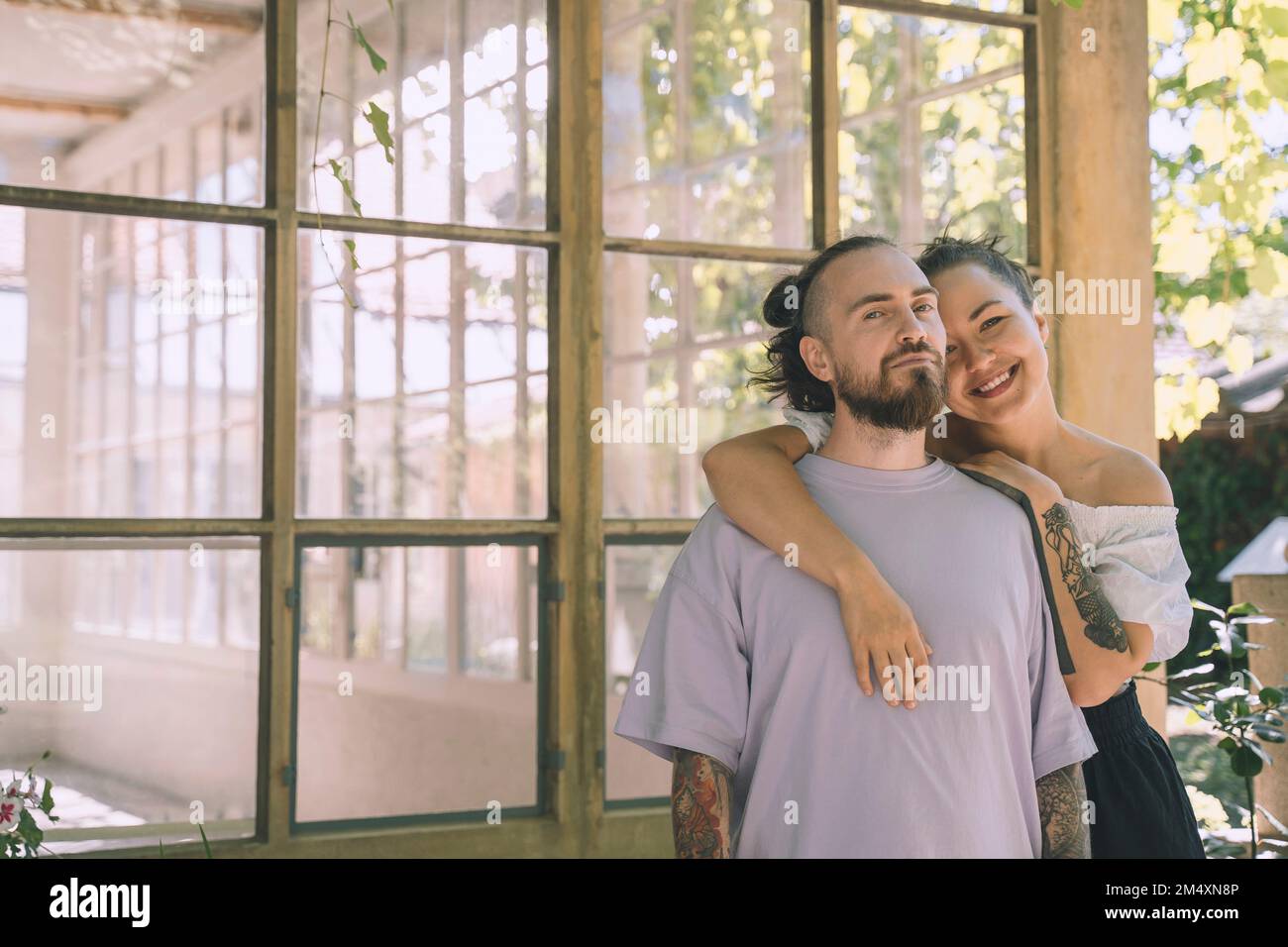 Smiling woman leaning on hipster boyfriend's shoulder in front of window Stock Photo