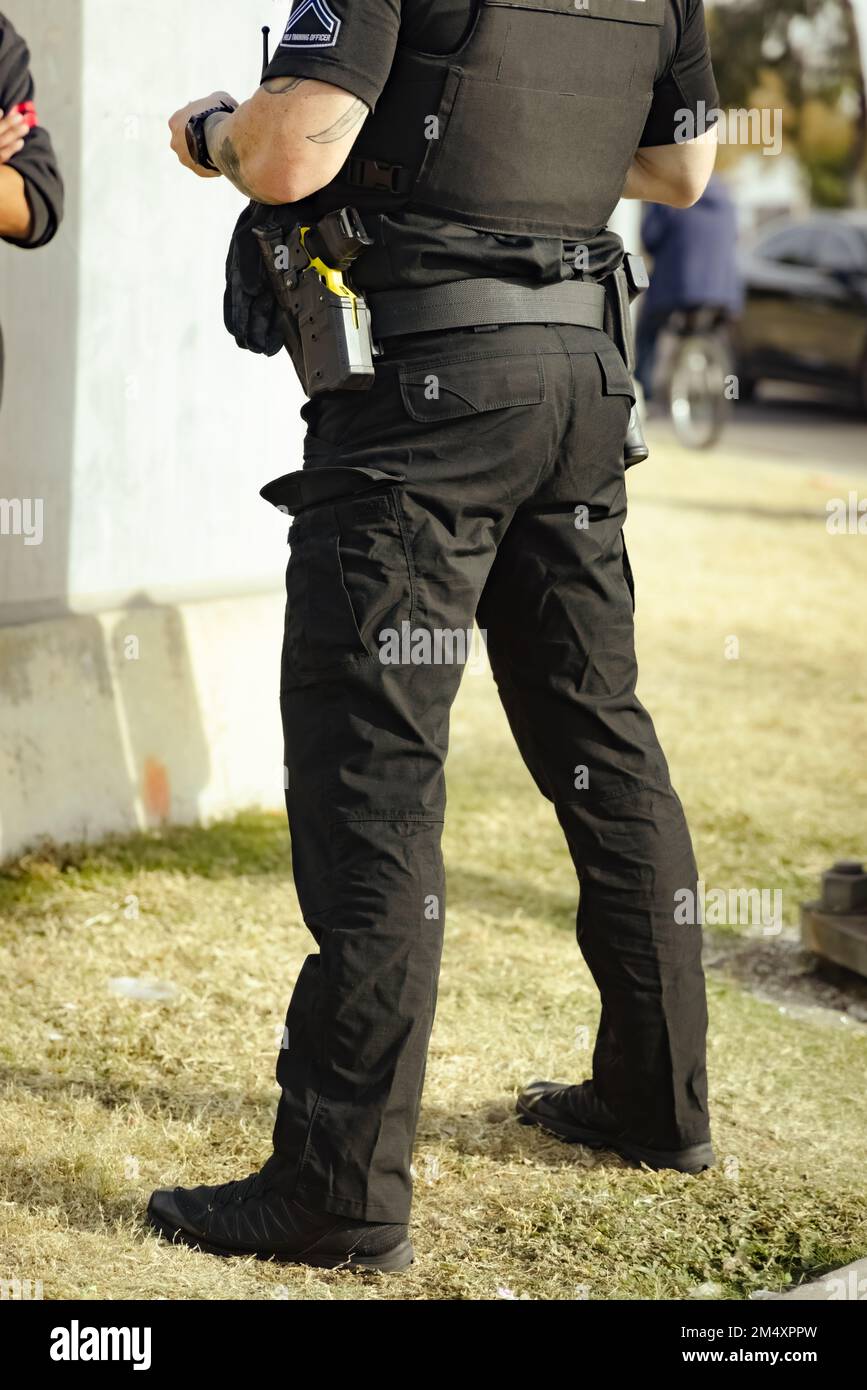 White male police officer cop standing on grass in a park during the day talking with person. Cop holding something in hand. Wearing bullet proof jack Stock Photo