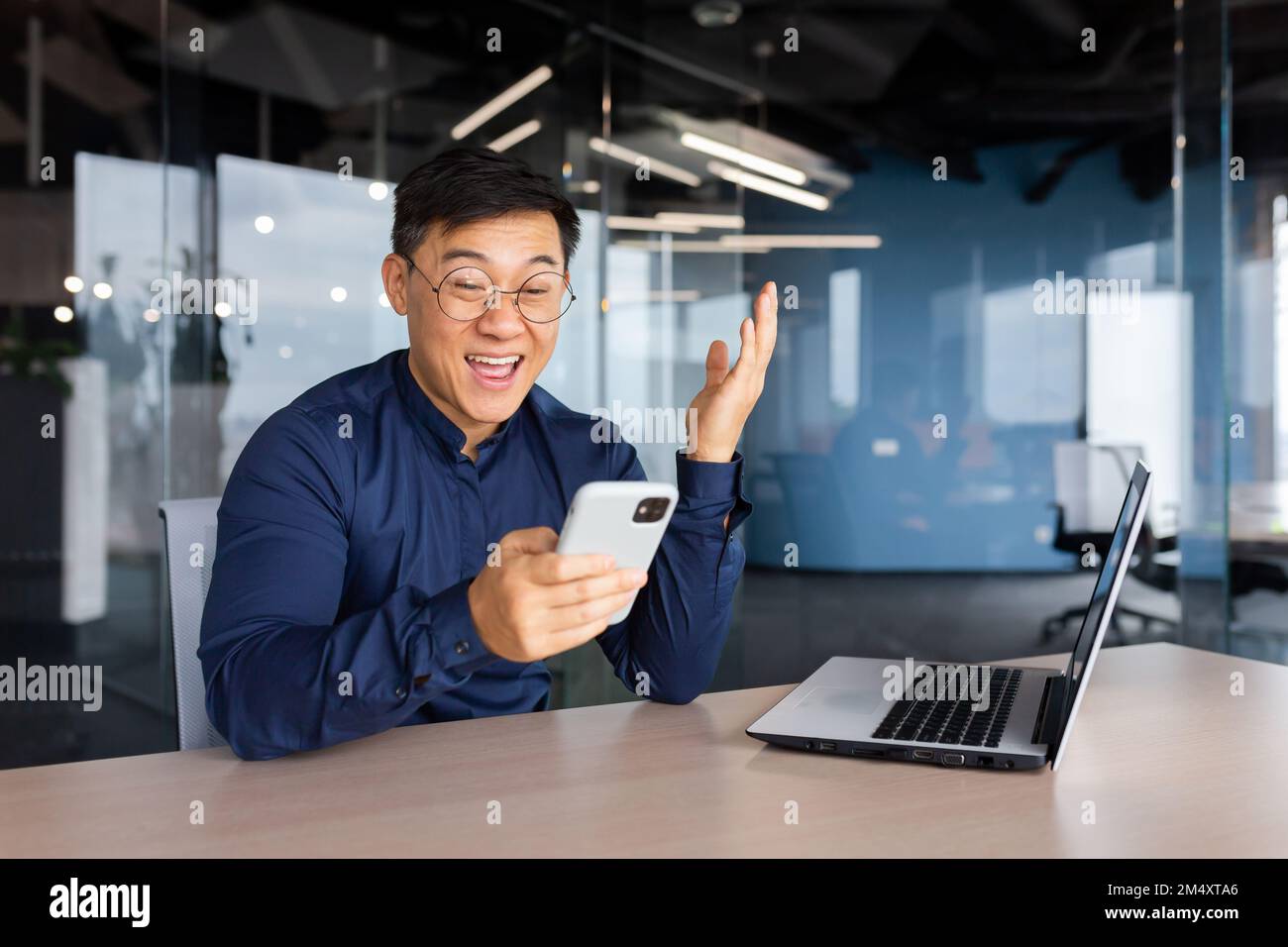 Happy young man businessman, freelancer sitting in the office at the table, holding the phone in his hands. Gladly reads the message, good news. Stock Photo