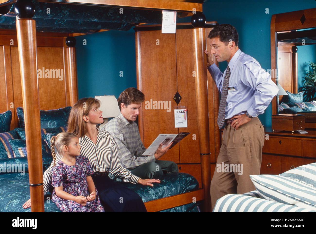 Young family, husband, wife and daughter sitting on a bed in a furniture store, speaking to the salesman Stock Photo
