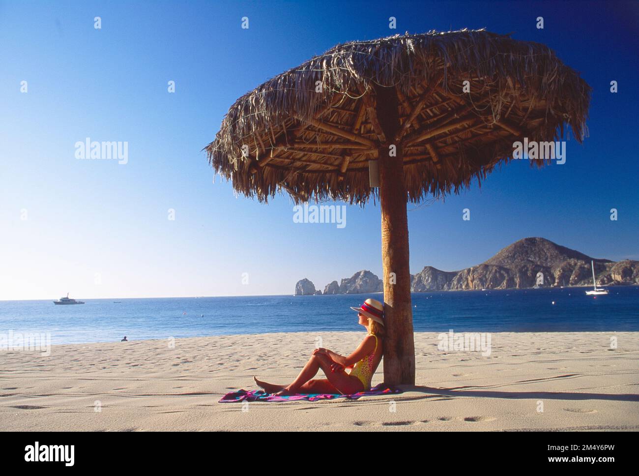 Younger woman. in a bathing suit and a hat sitting on the beach, leaning against a man-made shade device with a view of the ocean and rocky coastline Stock Photo