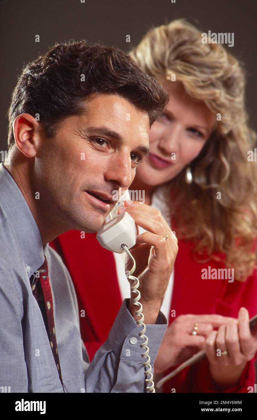 Office situation with businessman on the phone while female secretary is taking notes in the background Stock Photo