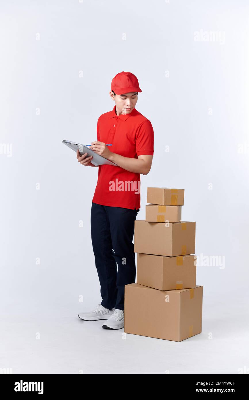 Cheerful professional male mover in casual wear making notes, holding clipboard, standing near a stack of cardboard boxes while doing home relocation. Stock Photo