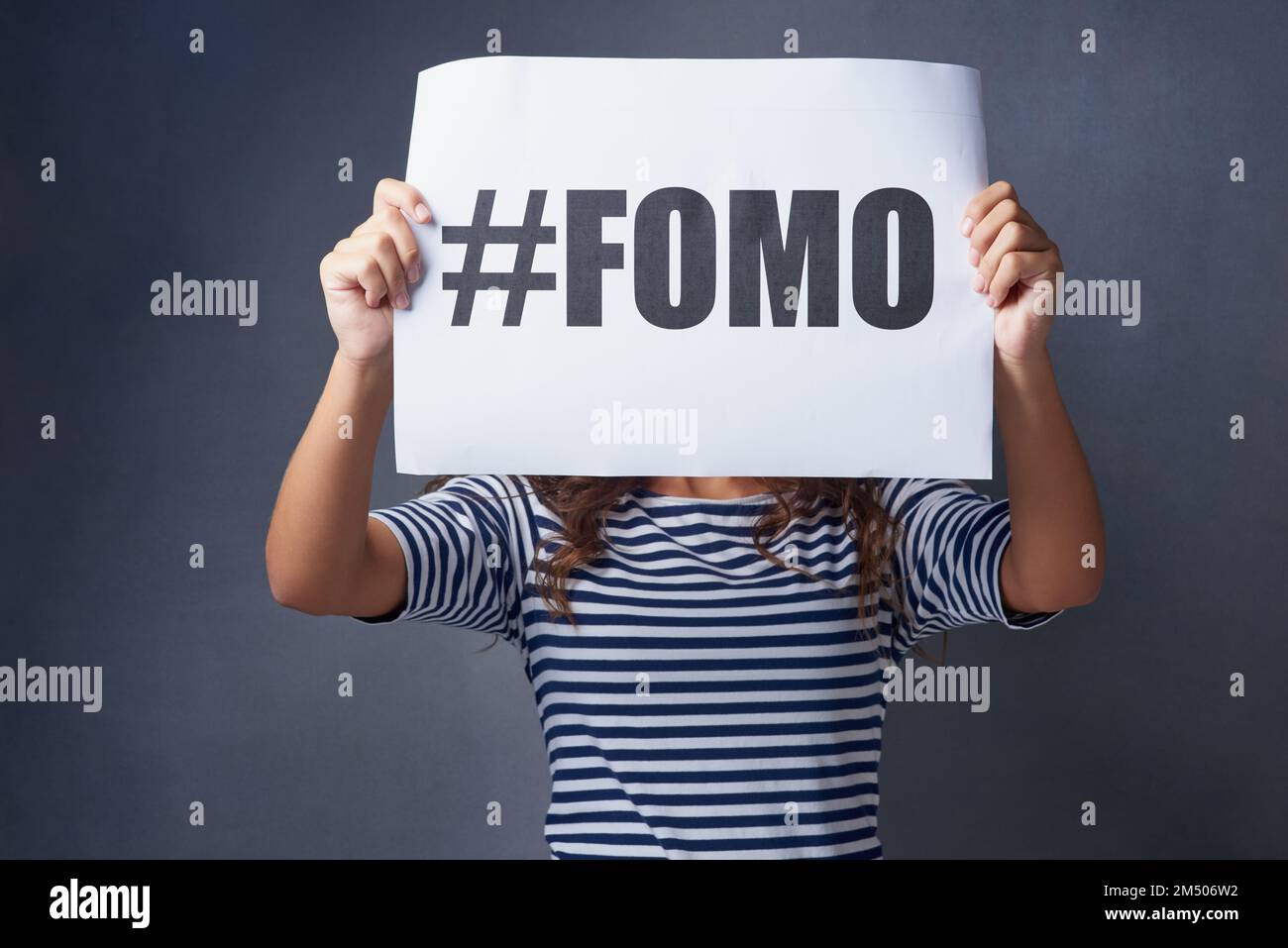 I feel so excluded. Studio shot of a young woman holding a sign with FOMO printed on it against a gray background. Stock Photo