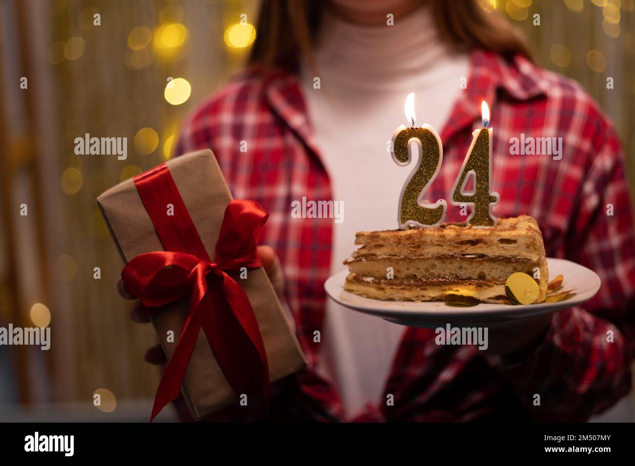 Young woman holding plate with tasty birthday cake and gift against defocused lights. 24th birthday. Copy space Stock Photo