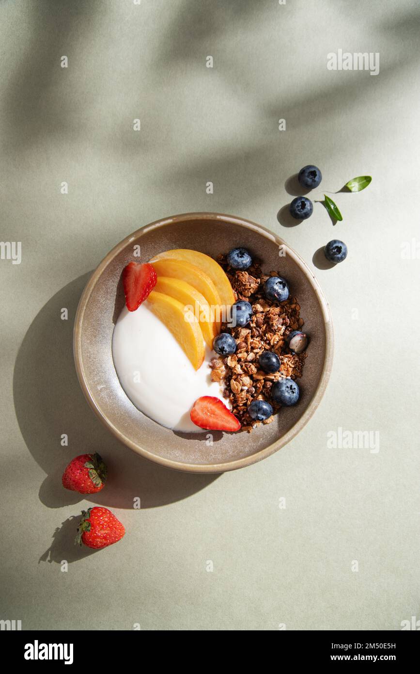 Delicious natural yogurt with homemade granola; peach, berries  in a bowl on a green background with morning shadows. Healthy and nutritious breakfast Stock Photo