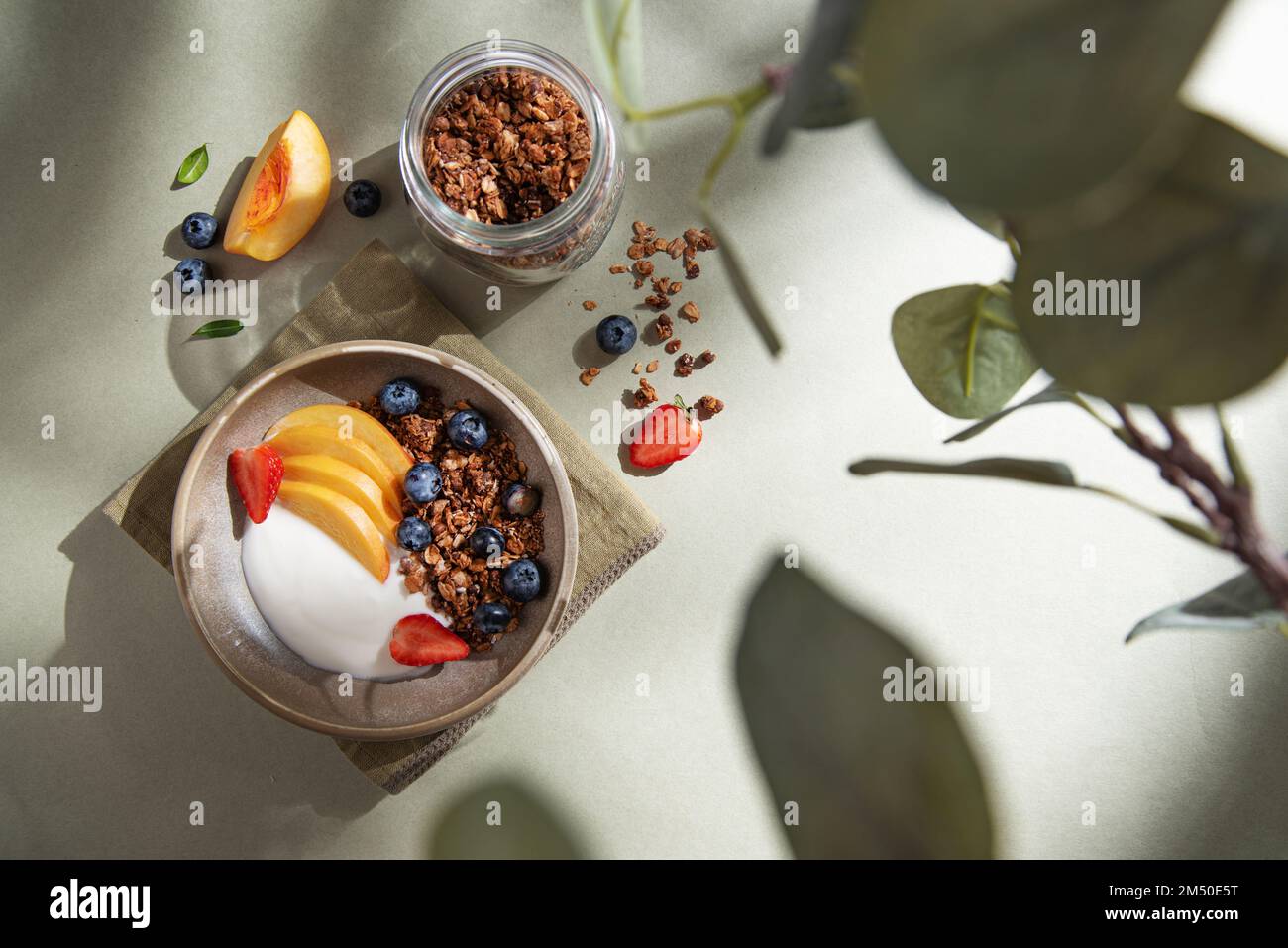 Delicious natural yogurt with homemade granola, berries,  peach, in a bowl and jar with muesli on a green background morning shadows. Healthy and nutr Stock Photo