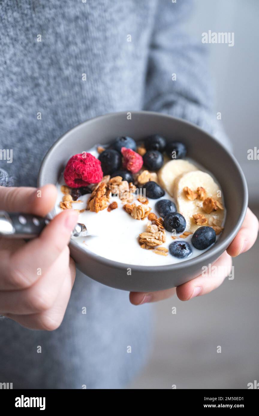 a young girl in a gray sweater is holding a bowl of vegetarian yogurt, granola and fresh blueberry and banana on the kitchen close up. The concept of Stock Photo