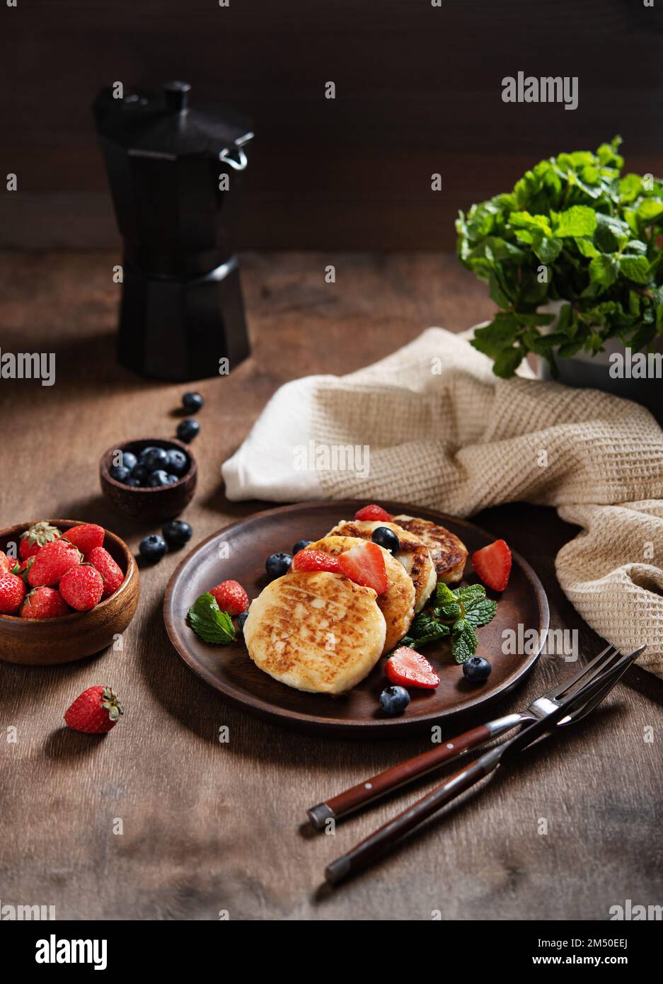 Cottage cheese pancakes or syrniki  with mint, strawberries and blueberries on a dark wooden table with a black coffee pot. Concept dark and moody hea Stock Photo