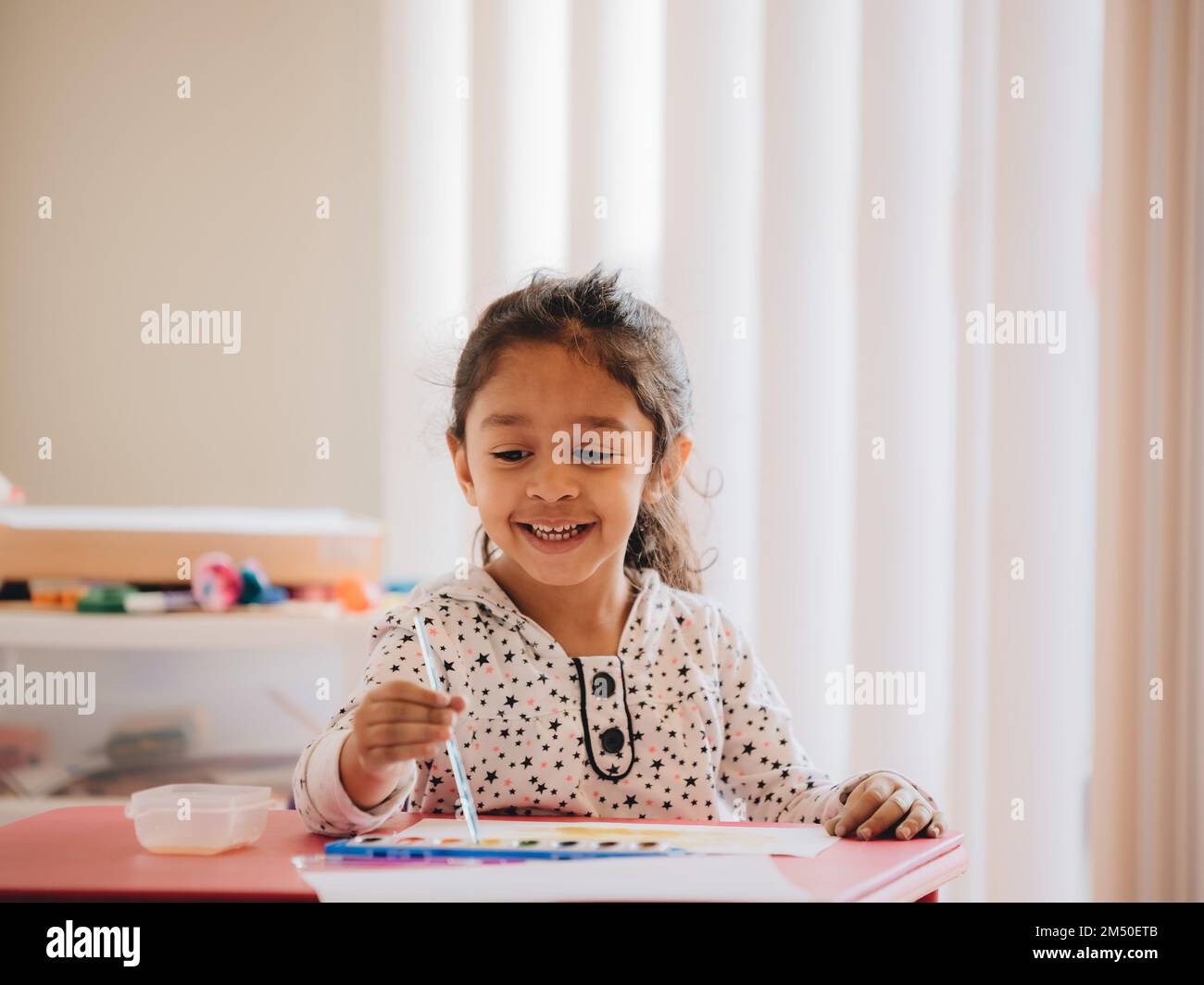 A cute preschool girl at home painting with watercolors on a piece of ...