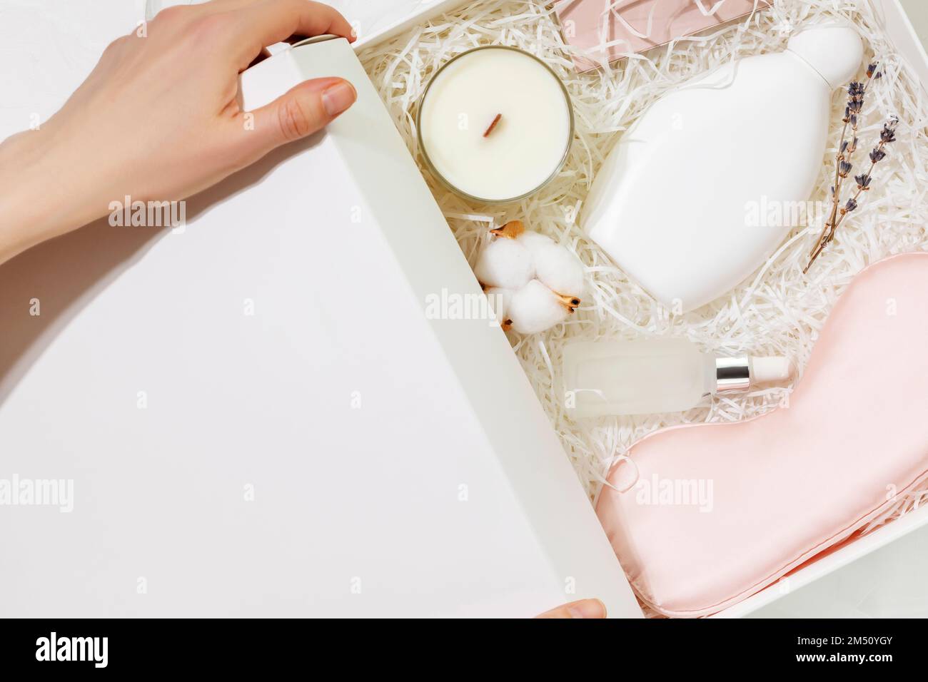 Female hands hold a white gift box with sleep care set. Preparing self care package, craft gift box with mask for sleep, aroma candle and bubble bath. Stock Photo