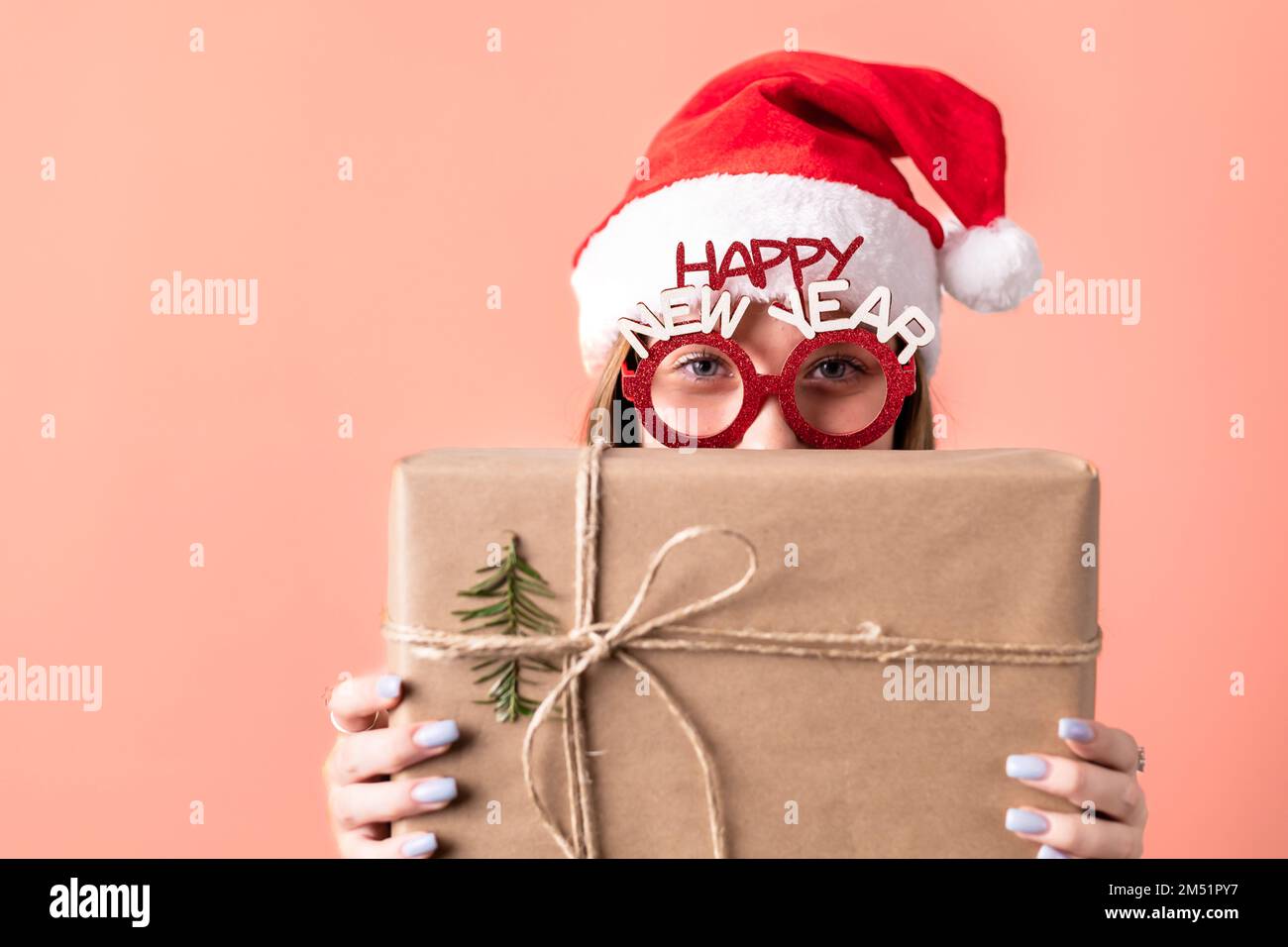 Beautiful, young, blonde girl with Santa hat and prop glasses isolated on red background, peaking behind a gift box. Copy space Stock Photo