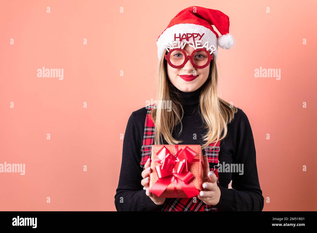 Beautiful, young, blonde girl with Santa hat and prop glasses isolated on red background, holding a gift box. Copy space Stock Photo
