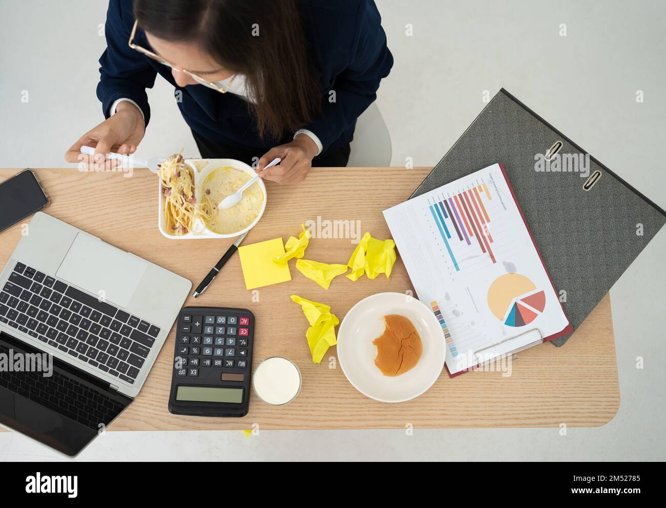 Top view of busy and tired businesswoman eating spaghetti for lunch at the Desk office and working to deliver financial statements to boss. Overworked Stock Photo