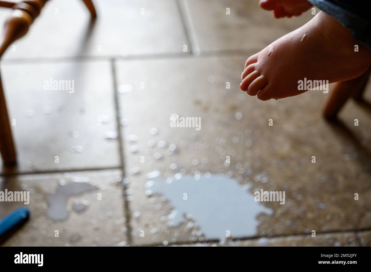 toddler foot suspended above spilled milk, crumbs, and a plastic fork on a brown tile floor  Stock Photo