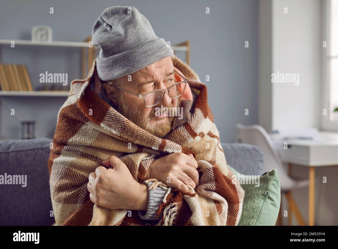 Senior man wearing warm clothes and blanket while freezing at home in cold winter Stock Photo