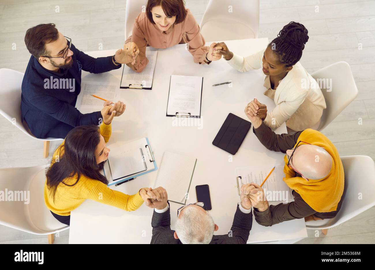 Diverse team of happy business people sitting around an office table and holding hands Stock Photo