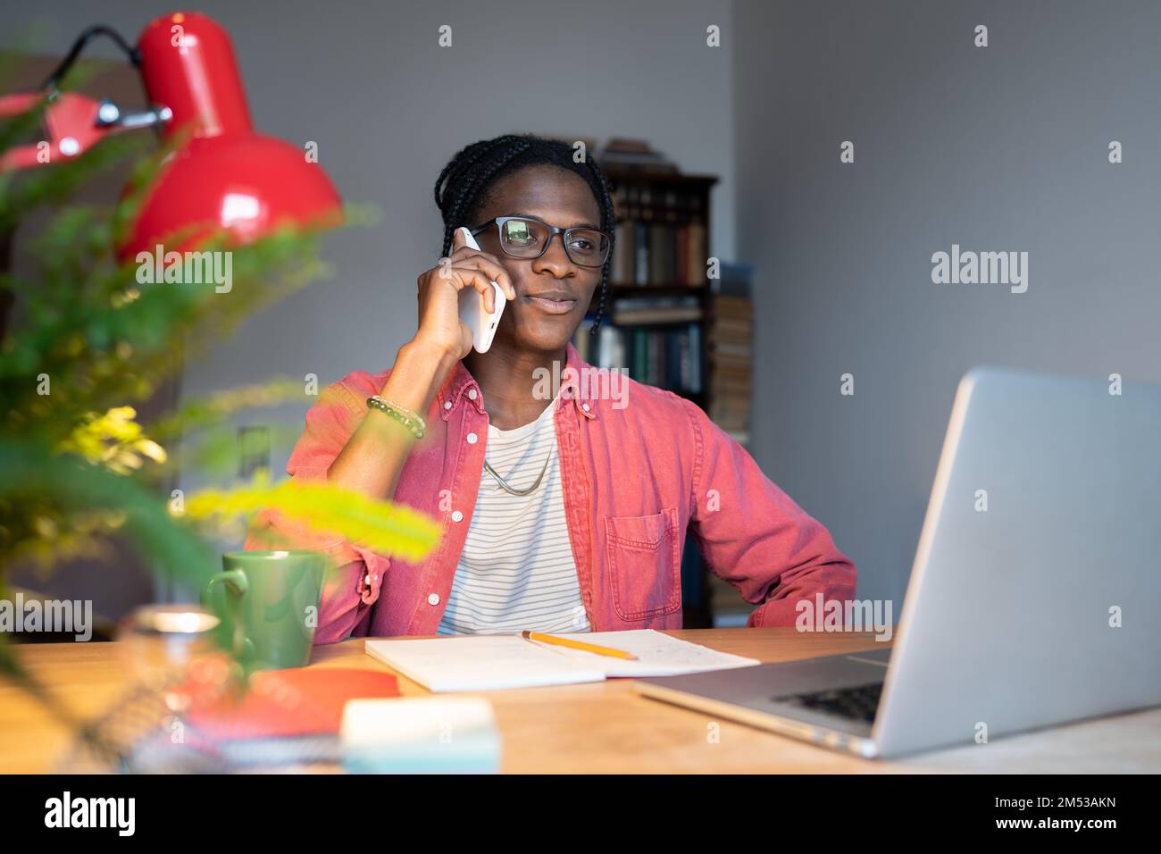Smiling African guy talking on phone while sitting at desk with laptop, distracted from online study Stock Photo
