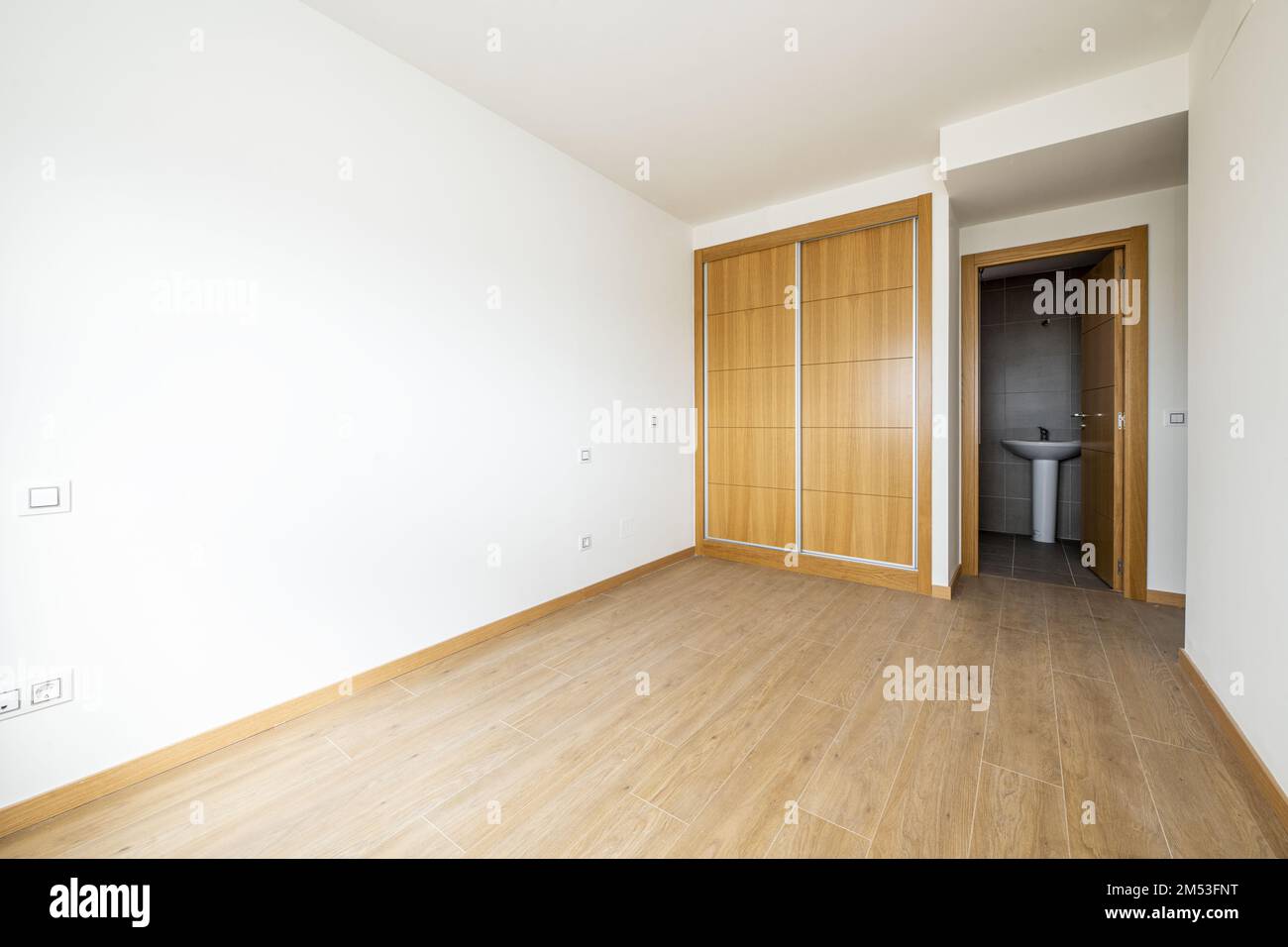 bedroom with a two-section built-in wardrobe with sliding oak doors and wood-like stoneware floors and access to an en-suite bathroom Stock Photo