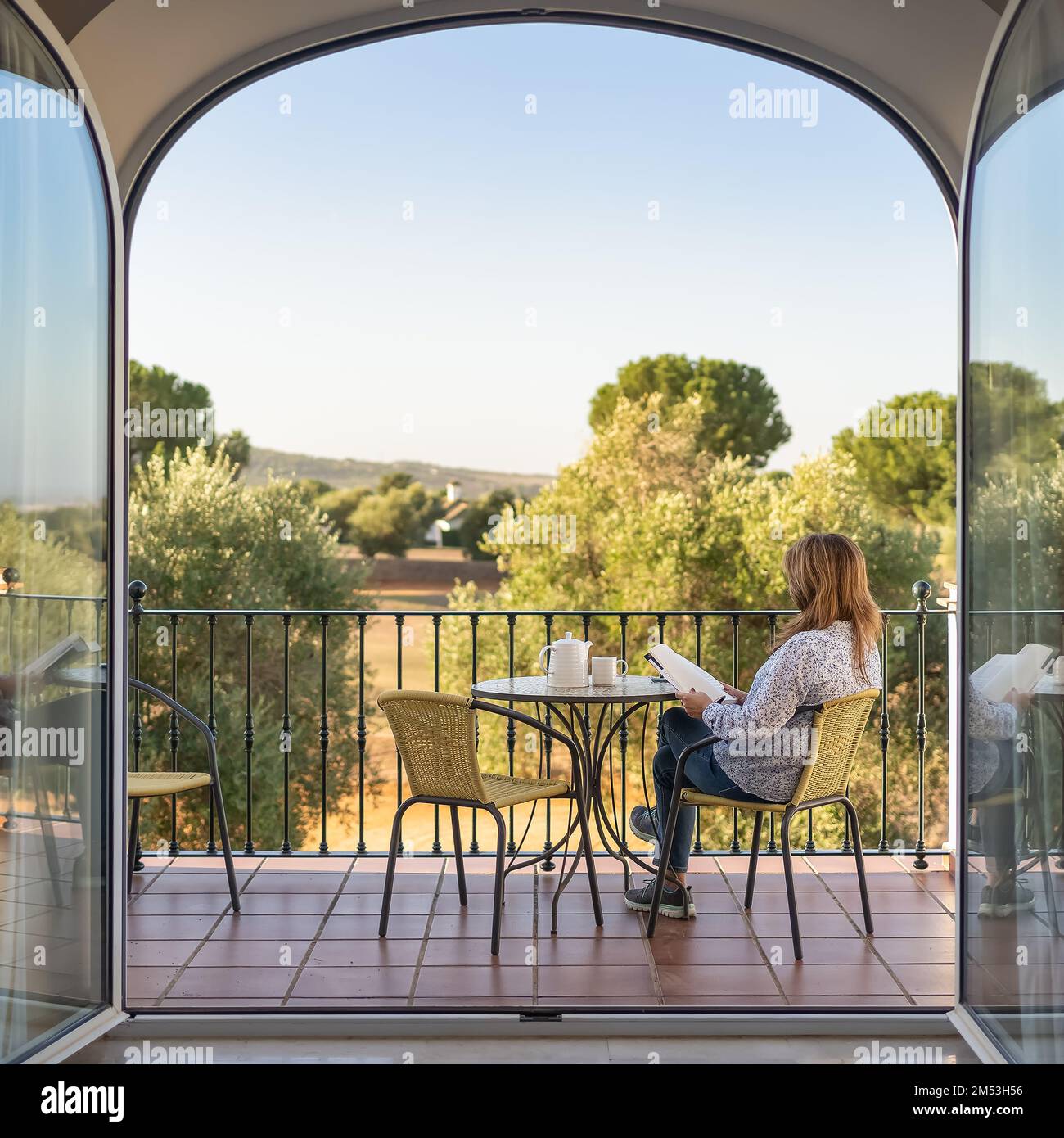 Woman reading a book on the terrace of her room with large glass doors and views to the outside of a large garden. Stock Photo