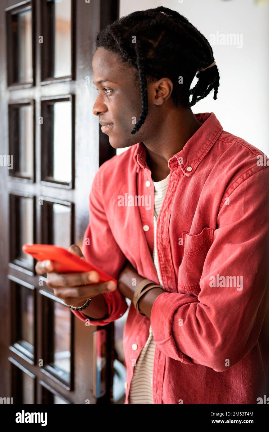 Thoughtful casual African American man with mobile phone stands indoors using messaging apps Stock Photo