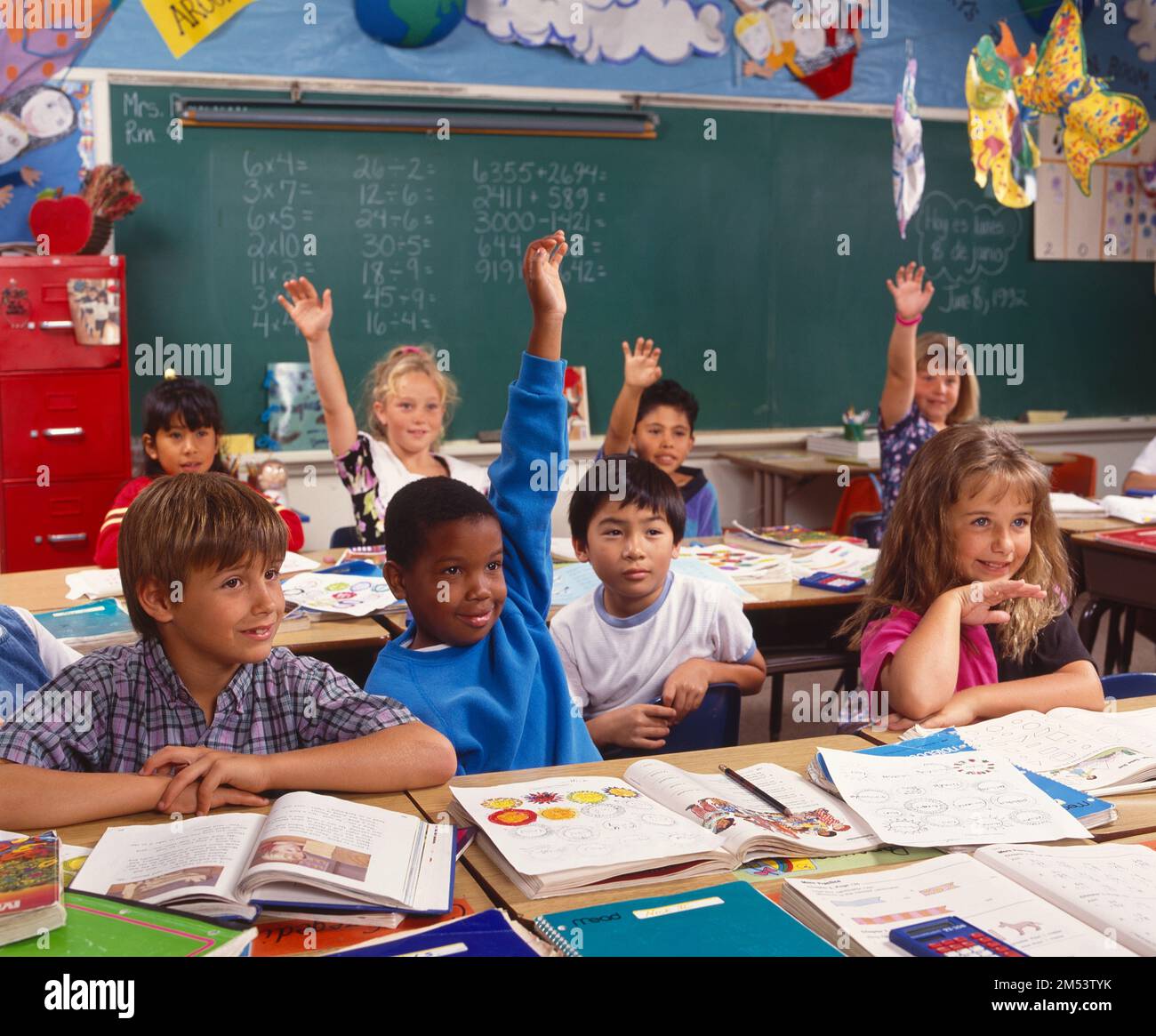 Children in a classroom, with their arms raised to be recognized by the ...