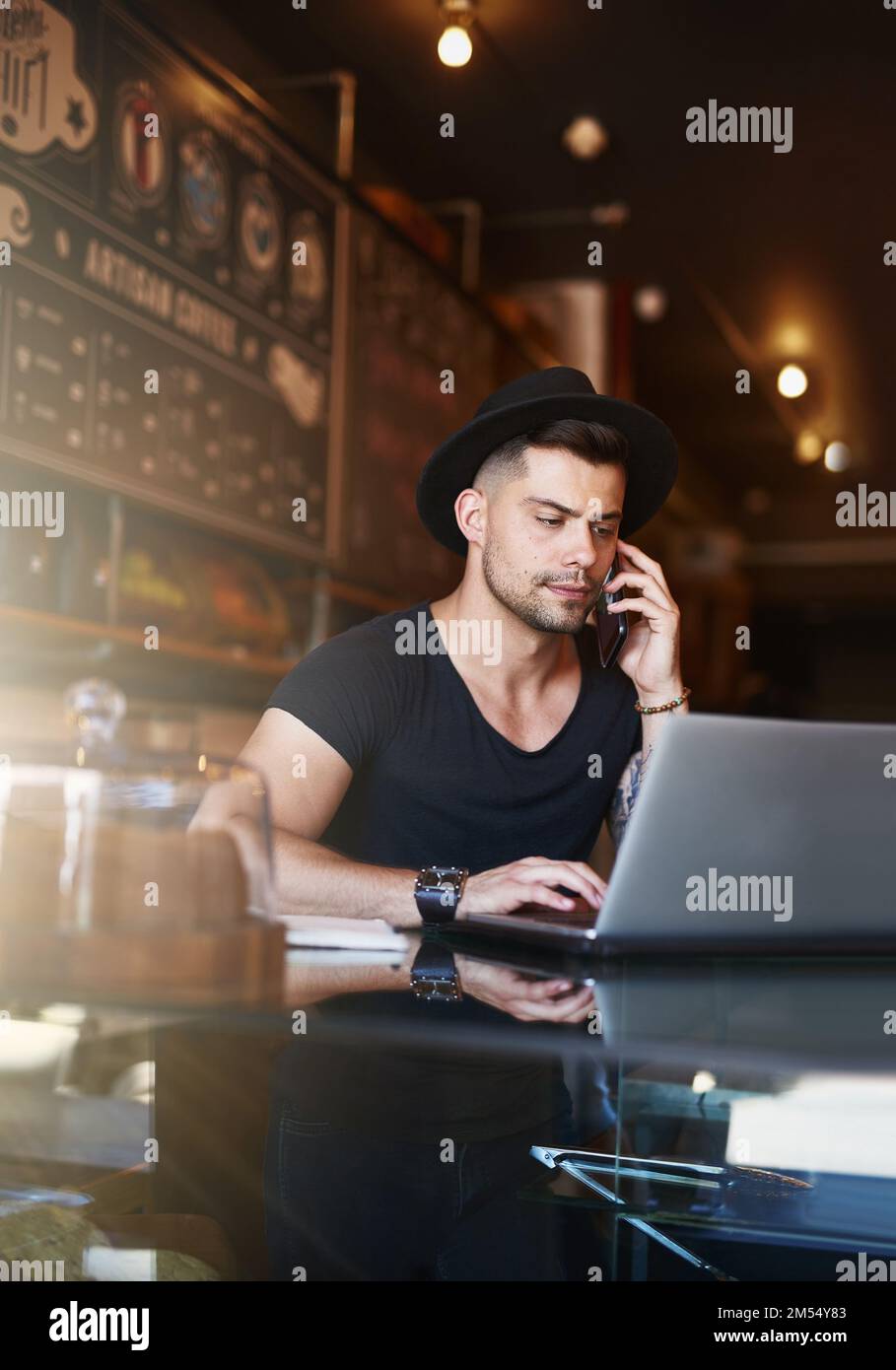 The hard work responsible for his cafes success. a young man using a phone and laptop while working in a coffee shop. Stock Photo