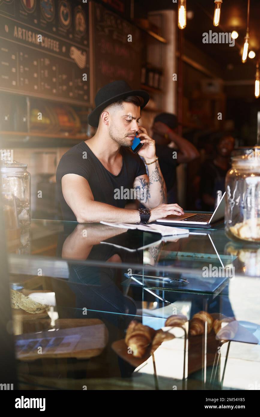 Fully involved in managing his coffee shop. a young man using a phone and laptop while working in a coffee shop. Stock Photo
