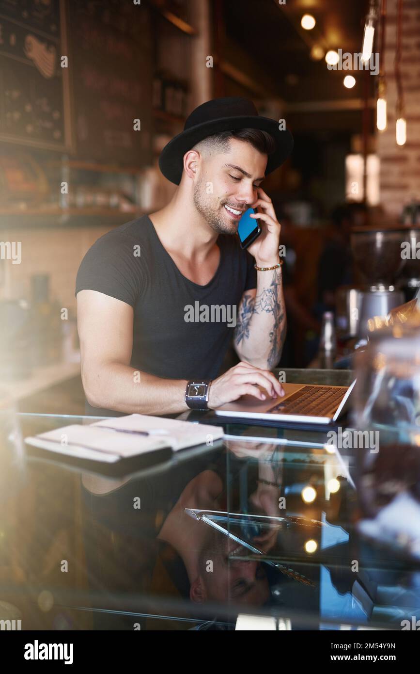 The coffee business is booming. a young man using a phone and laptop while working in a coffee shop. Stock Photo