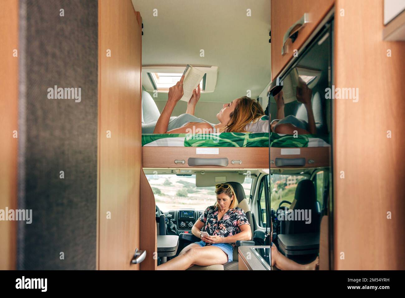 Woman lying on camper van bunk bed reading book while her friend looking mobile Stock Photo